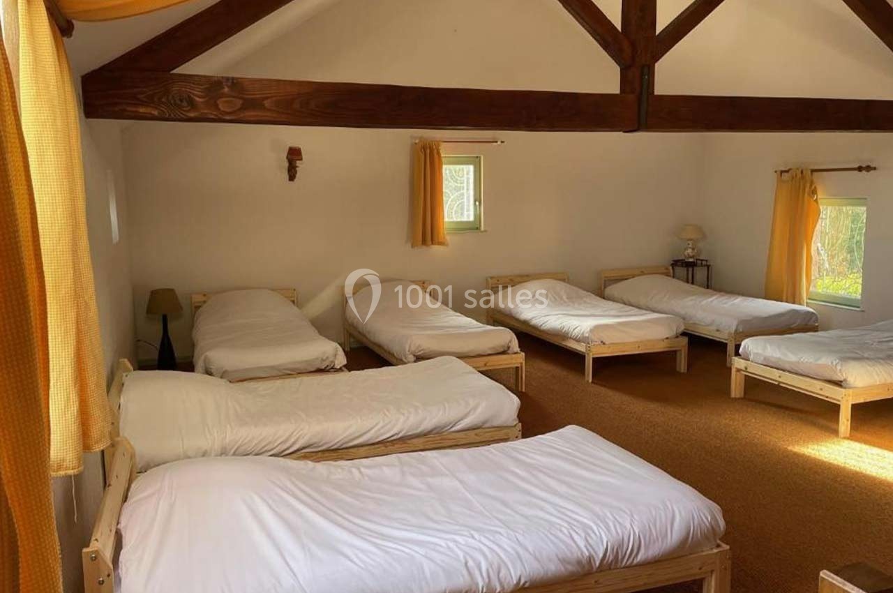 Dortoir lumineux avec six lits simples en bois, draps blancs, poutres apparentes et fenêtres avec rideaux jaunes.