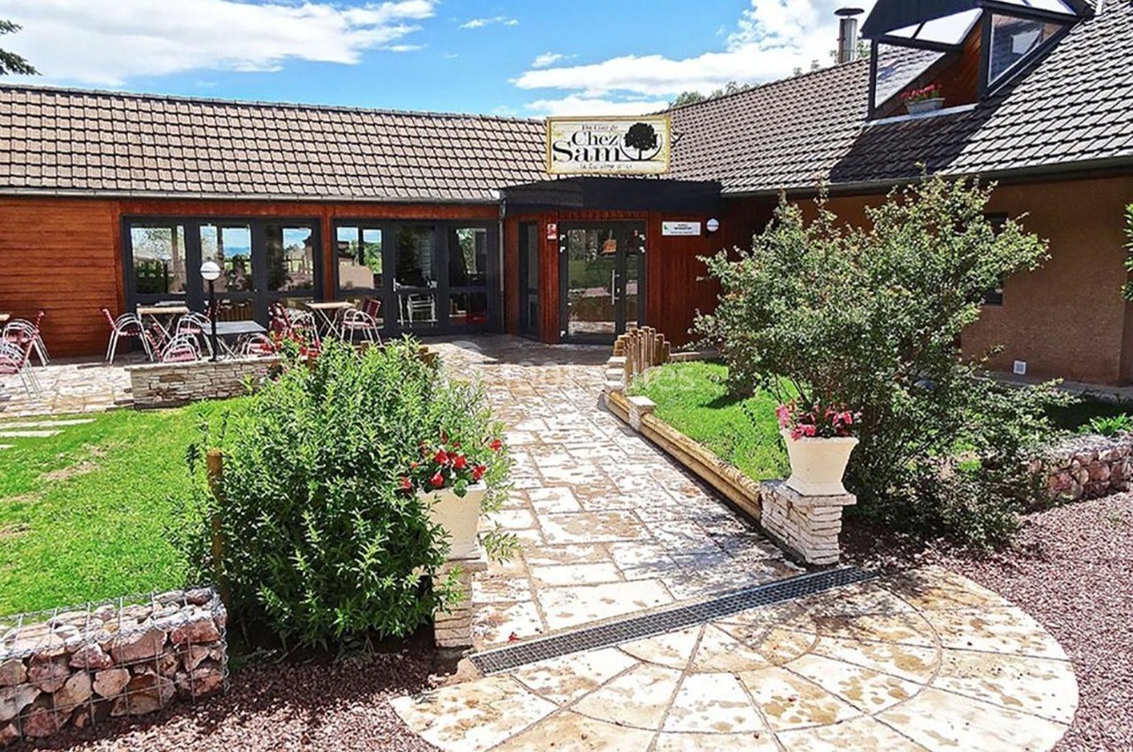 Entrée d'un restaurant avec terrasse, tables extérieures et végétation environnante sous un ciel dégagé.