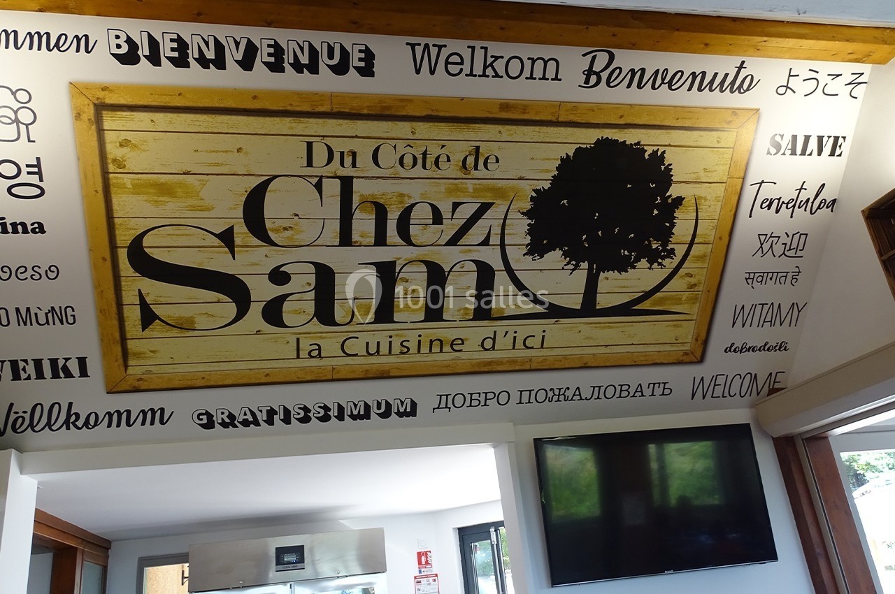 Panneau en bois avec l'inscription ’Du Côté de Chez Sam, la Cuisine d'ici’, entouré de mots ’Bienvenue’ en plusieurs langues.