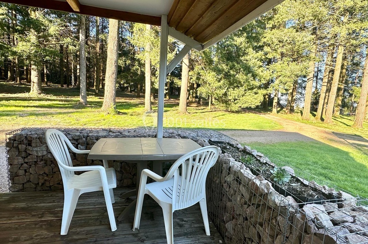 Terrasse en bois avec table et chaises blanches, entourée d'un muret en pierre, donnant sur une forêt ensoleillée.