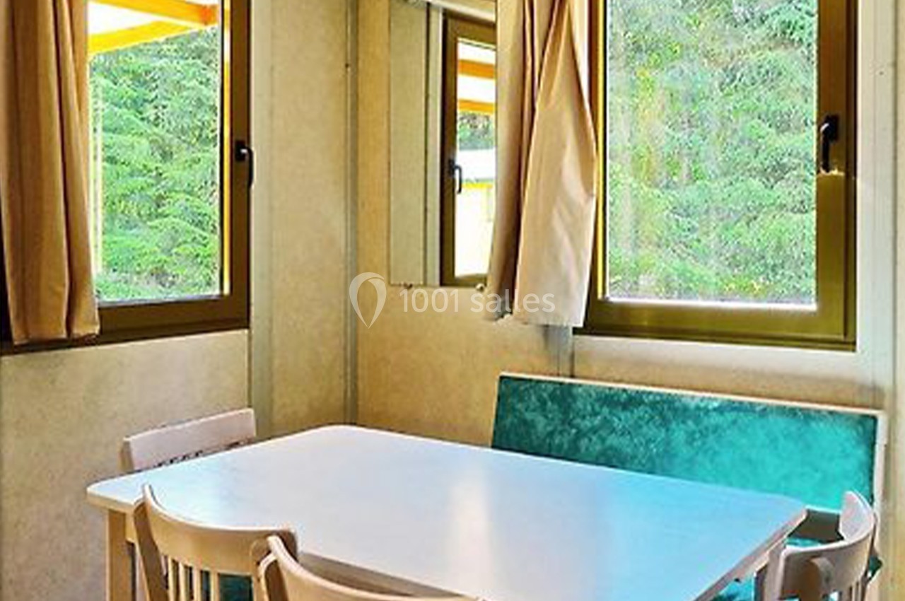 Salle à manger lumineuse avec table blanche, chaises en bois et banquette verte, entourée de fenêtres avec rideaux beiges.