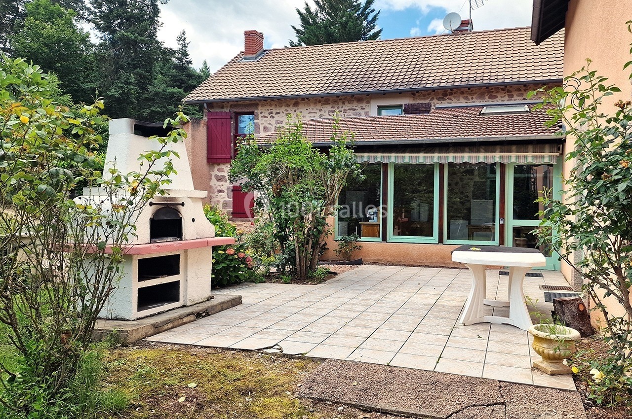 Terrasse carrelée avec barbecue en pierre, table blanche et maison en pierre avec fenêtres et volets rouges en arrière-plan.