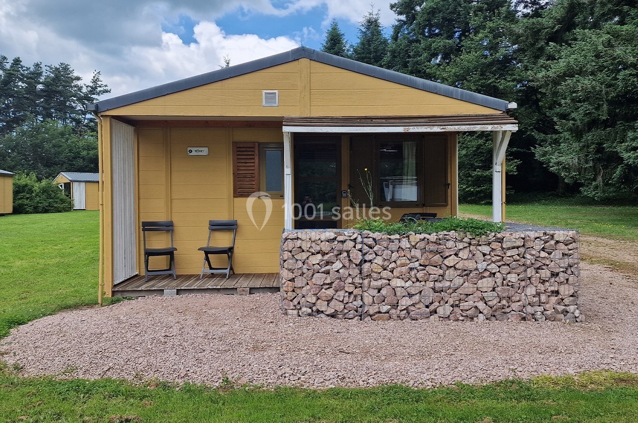 Chalet en bois jaune avec terrasse couverte, deux chaises, muret en pierres et pelouse environnante.