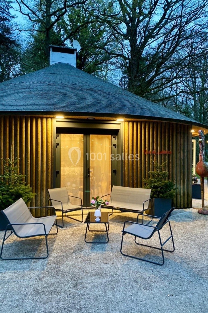 Espace extérieur avec fauteuils et table basse devant une cabane ronde éclairée, entourée d'arbres.