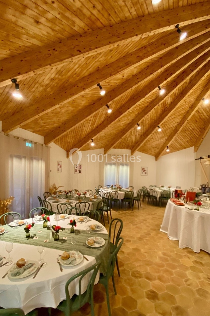 Salle de réception avec tables rondes dressées, décor rustique avec plafond en bois et lumière chaleureuse.