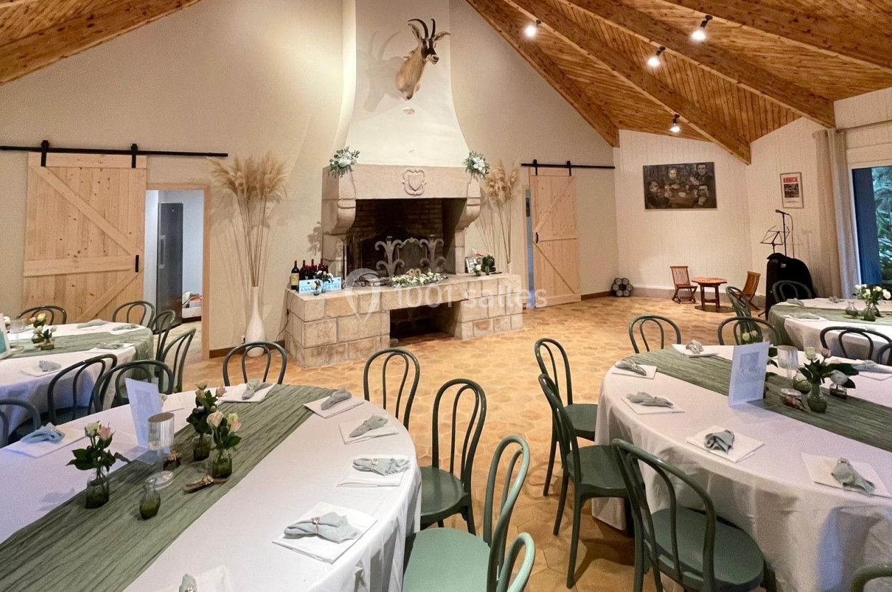 Salle décorée pour un événement, avec tables rondes dressées, chaises vertes et cheminée ornée sous un plafond en bois.