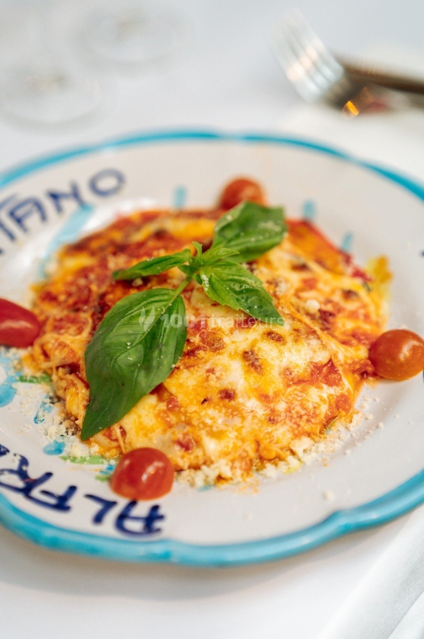 Assiette de lasagnes garnie de basilic frais et entourée de petites tomates cerises, servie sur une table dressée.