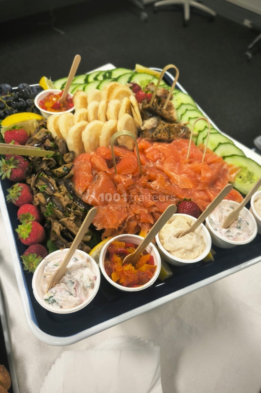 Plateau de saumon fumé avec légumes frais, fruits, dips variés et crackers, présenté sur une table.