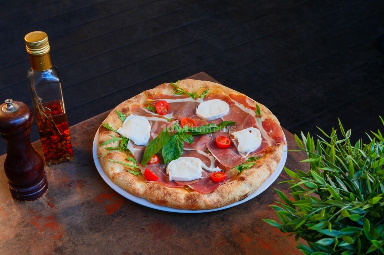 Pizza garnie de jambon cru, fromage, tomates cerises et roquette, posée sur une table avec des condiments et une plante.