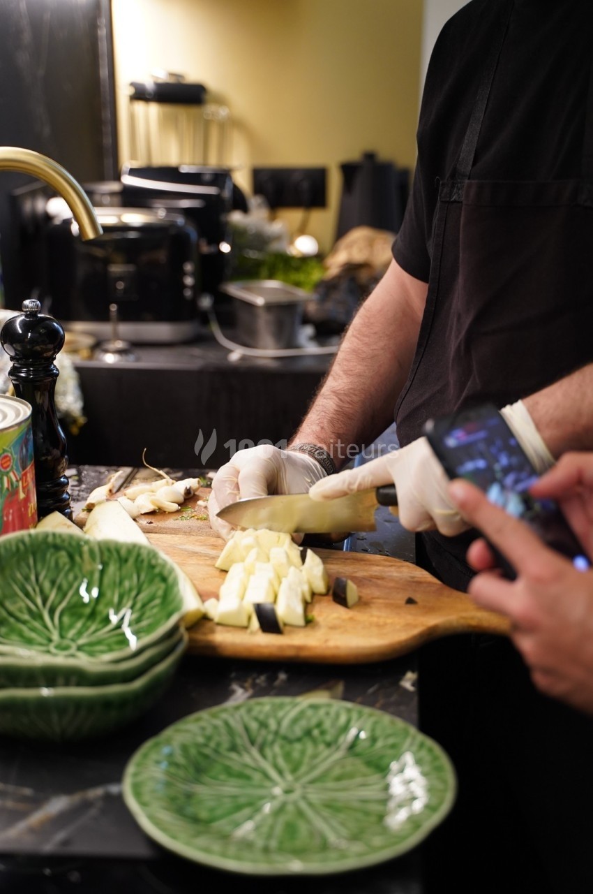 Un cuisinier découpe des légumes sur une planche en bois, avec des assiettes vertes en premier plan.