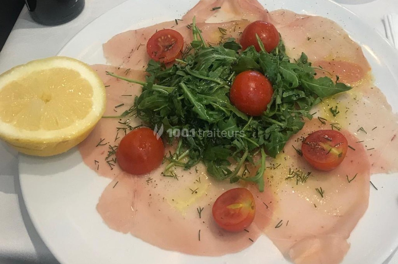 Assiette de carpaccio de poisson garnie de roquette, tomates cerises et herbes, accompagnée d’un demi-citron.