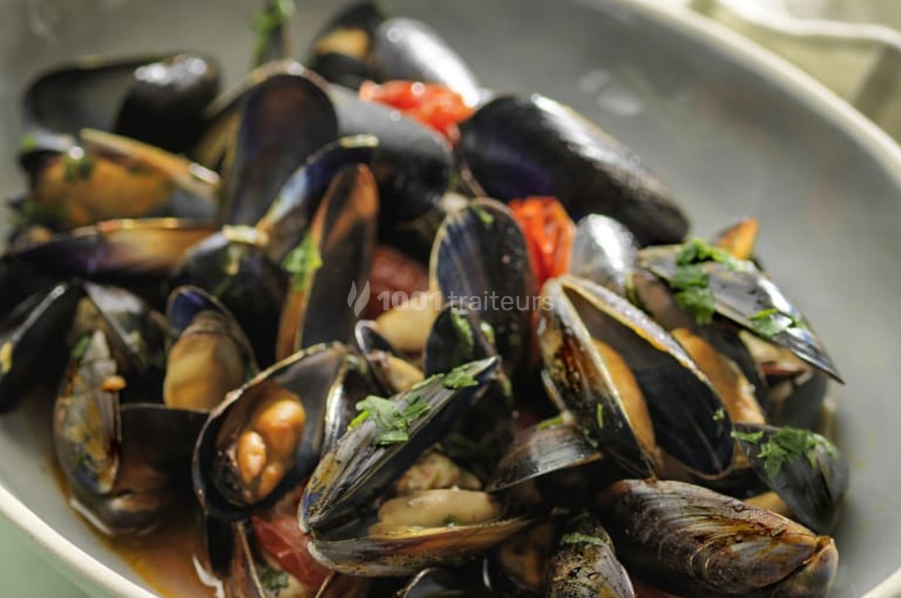 Assiette de moules cuisinées avec tomates, herbes fraîches et sauce, servie dans un plat creux.