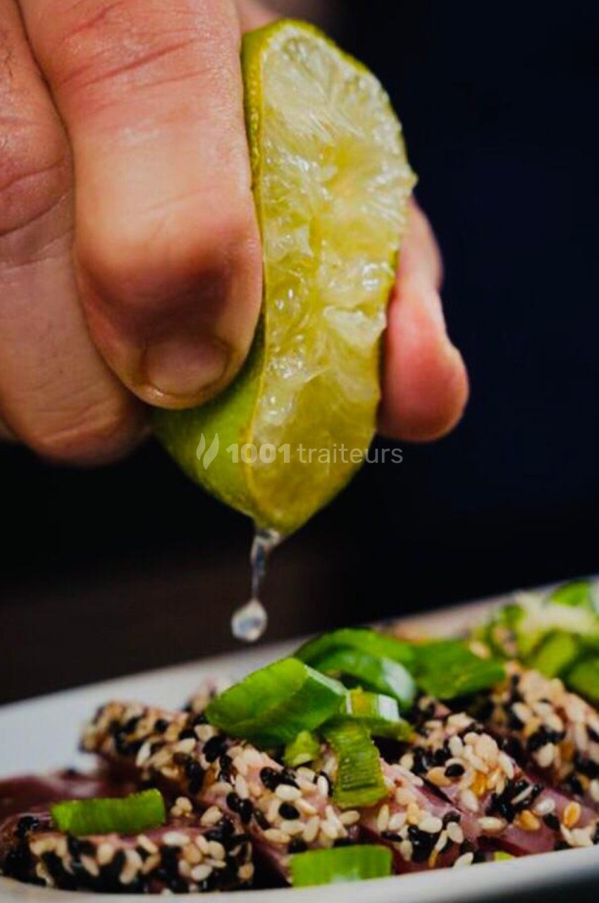 Une main presse un demi-citron vert au-dessus d'un plat garni de graines de sésame et de morceaux de légumes verts.