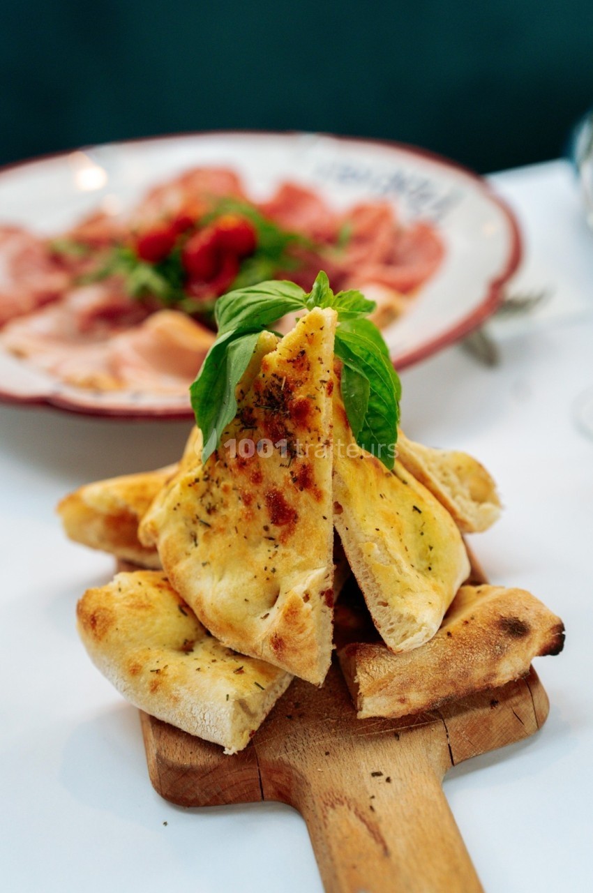 Tranches de pain plat doré garnies d'herbes, servies sur une planche en bois, avec un plat de charcuterie en arrière-plan.