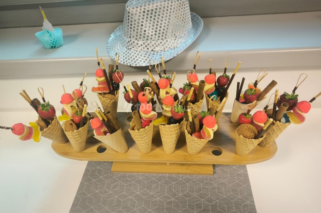Présentoir en bois avec des cornets garnis de fruits, bonbons et biscuits, posé sur une table près d'un chapeau argenté.