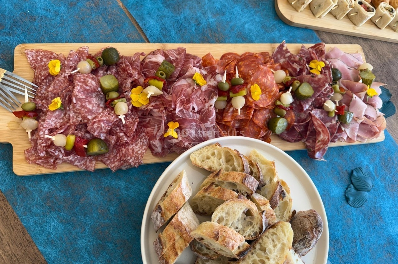 Plateau de charcuterie variée avec olives et fleurs comestibles, accompagné de tranches de pain sur une table bleue.