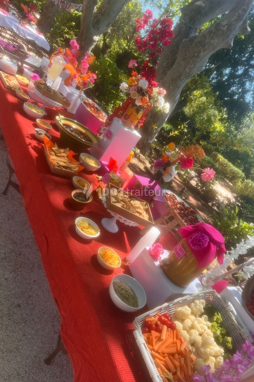 Table garnie de plats variés, sauces et légumes, disposée en extérieur sous des arbres et entourée de fleurs colorées.