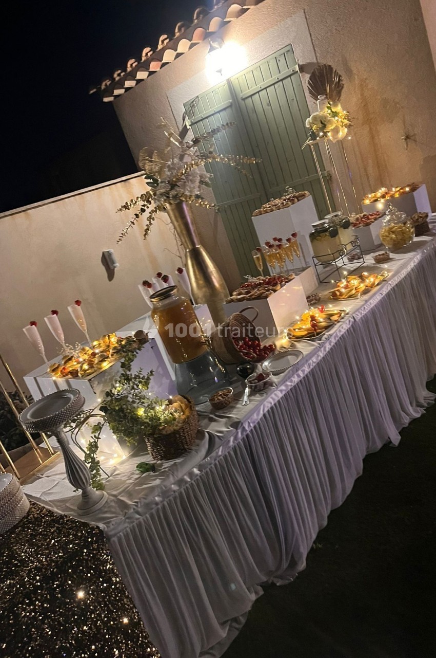 Table décorée pour un buffet en extérieur, éclairée par des guirlandes lumineuses, avec desserts et boissons disposés.