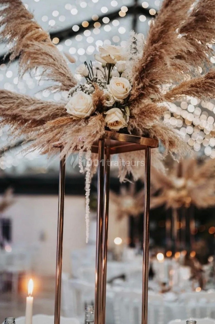 Centre de table avec des pampas, des roses blanches et des bougies, sous une verrière illuminée de guirlandes.