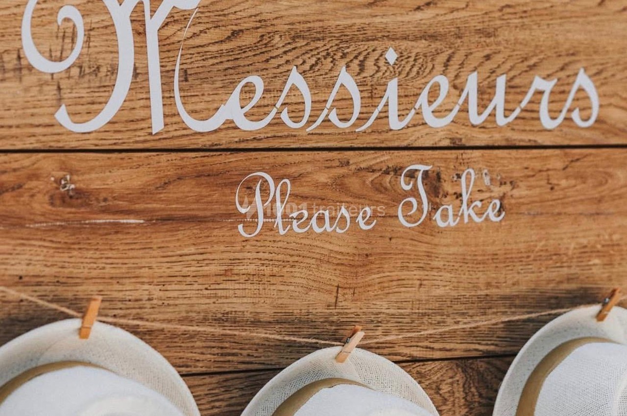 Panneau en bois avec l'inscription ’Messieurs, please take’ et plusieurs chapeaux blancs suspendus.
