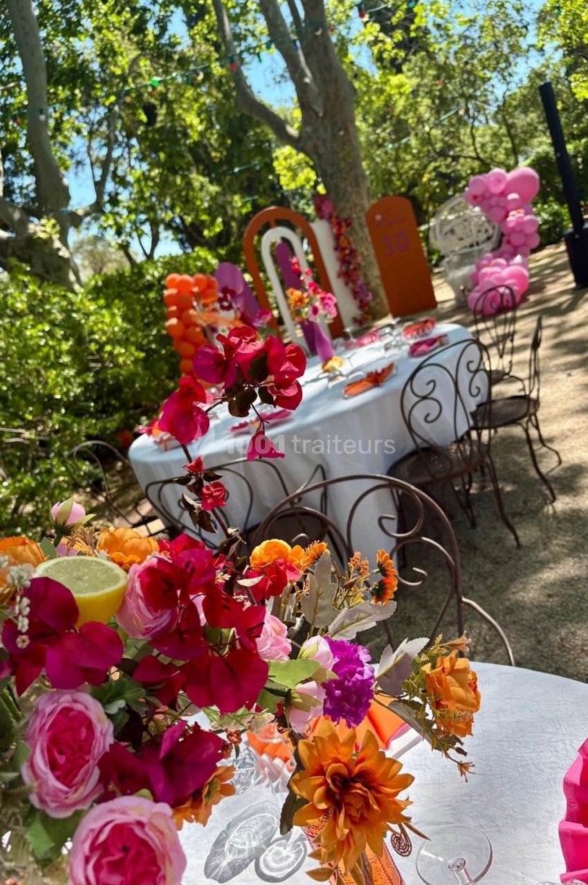 Tables décorées avec des fleurs colorées dans un jardin, entourées de ballons et d'arbres sous une lumière naturelle.