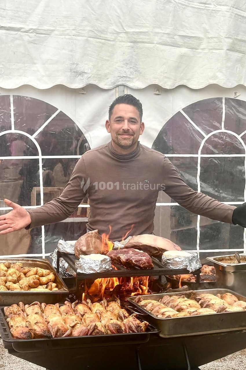 Homme souriant devant un barbecue chargé de viandes et pommes de terre, sous une tente blanche.