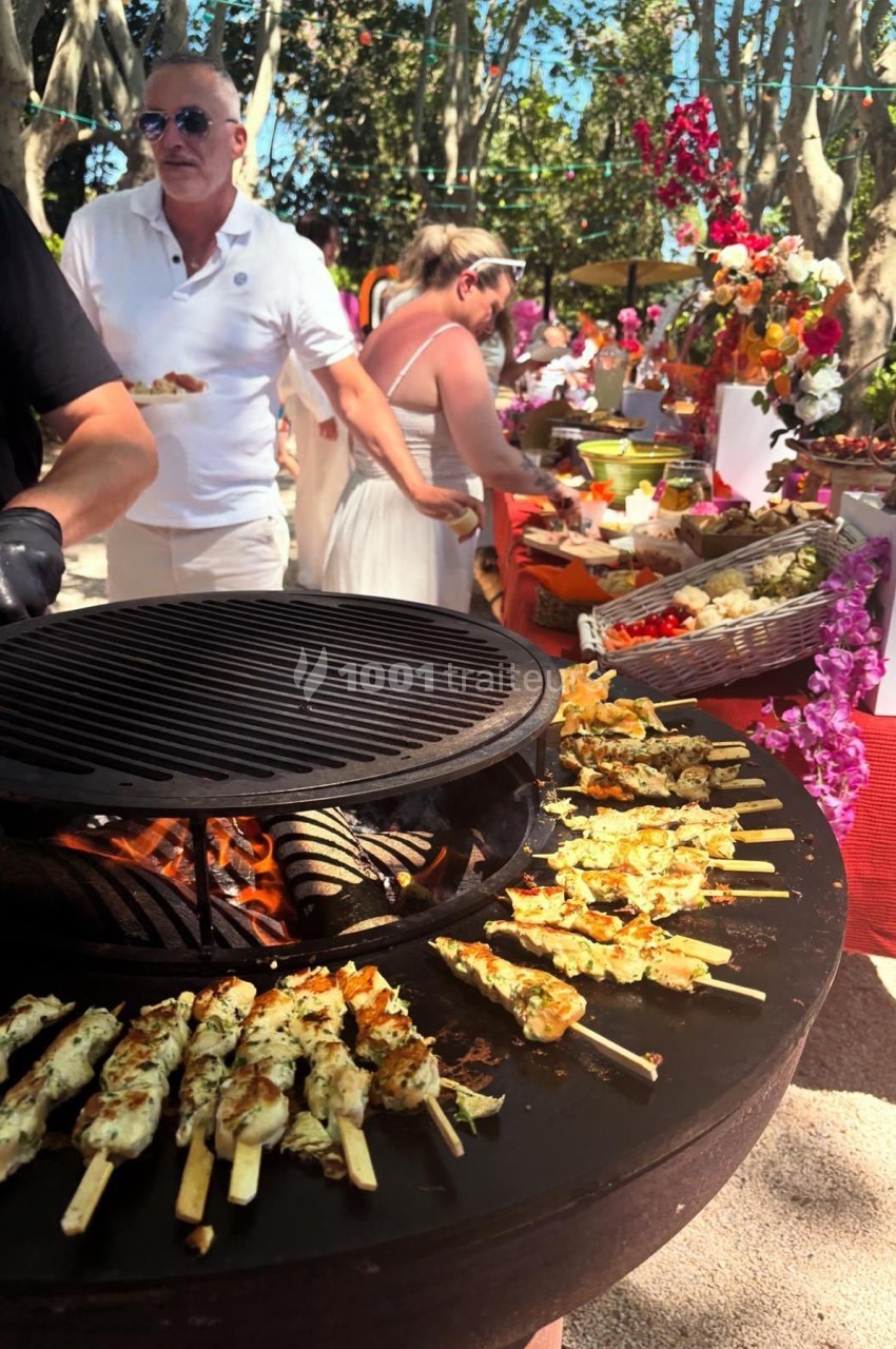 Grill circulaire avec brochettes en cuisson, buffet coloré en arrière-plan et personnes se servant de la nourriture.