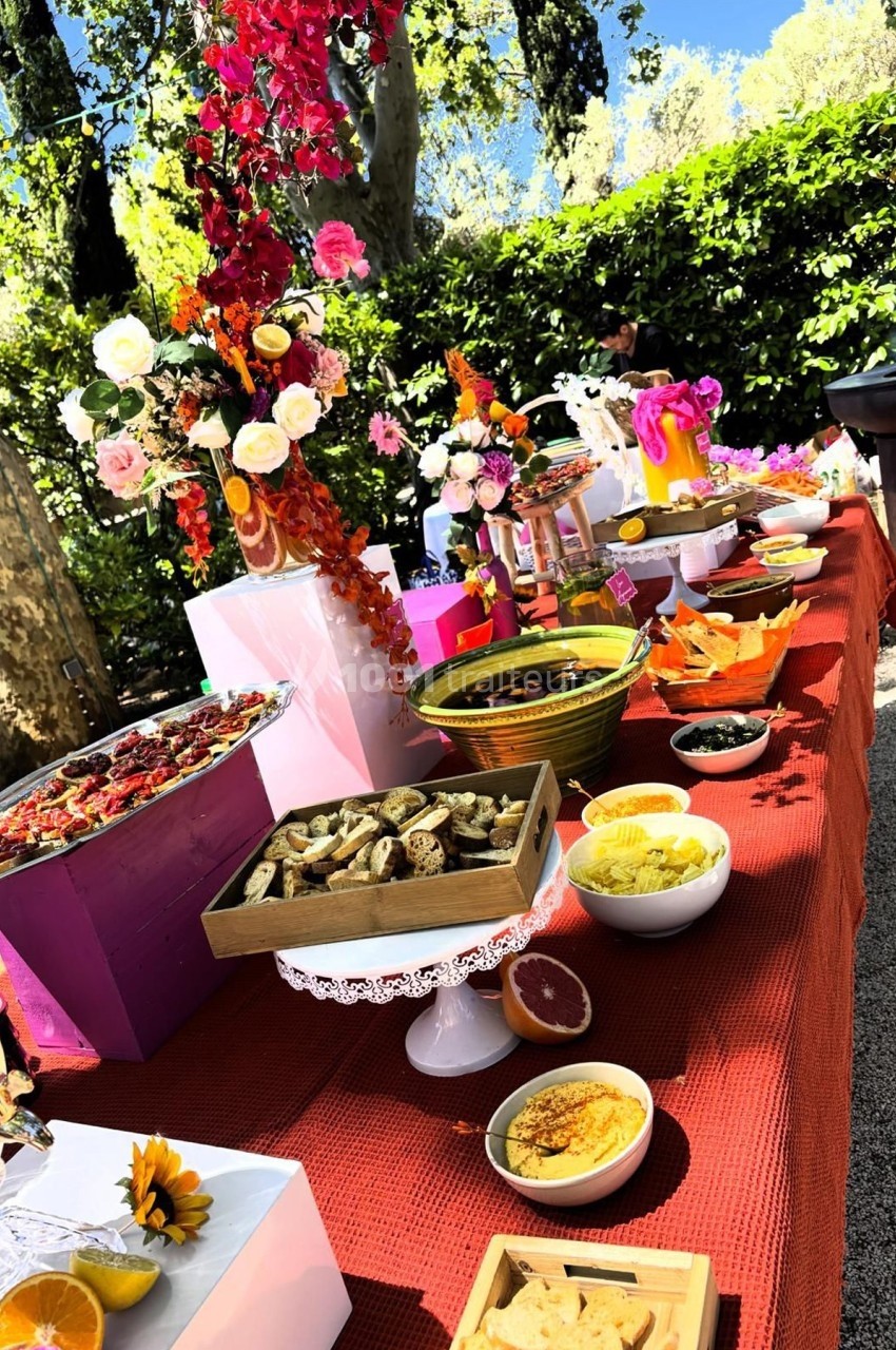 Table garnie de plats variés, fleurs colorées et décorations, disposée en extérieur sous un ciel ensoleillé.