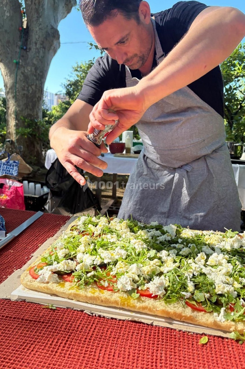 Un homme en tablier assaisonne une grande tarte salée garnie de légumes et fromage, dans un jardin ensoleillé.