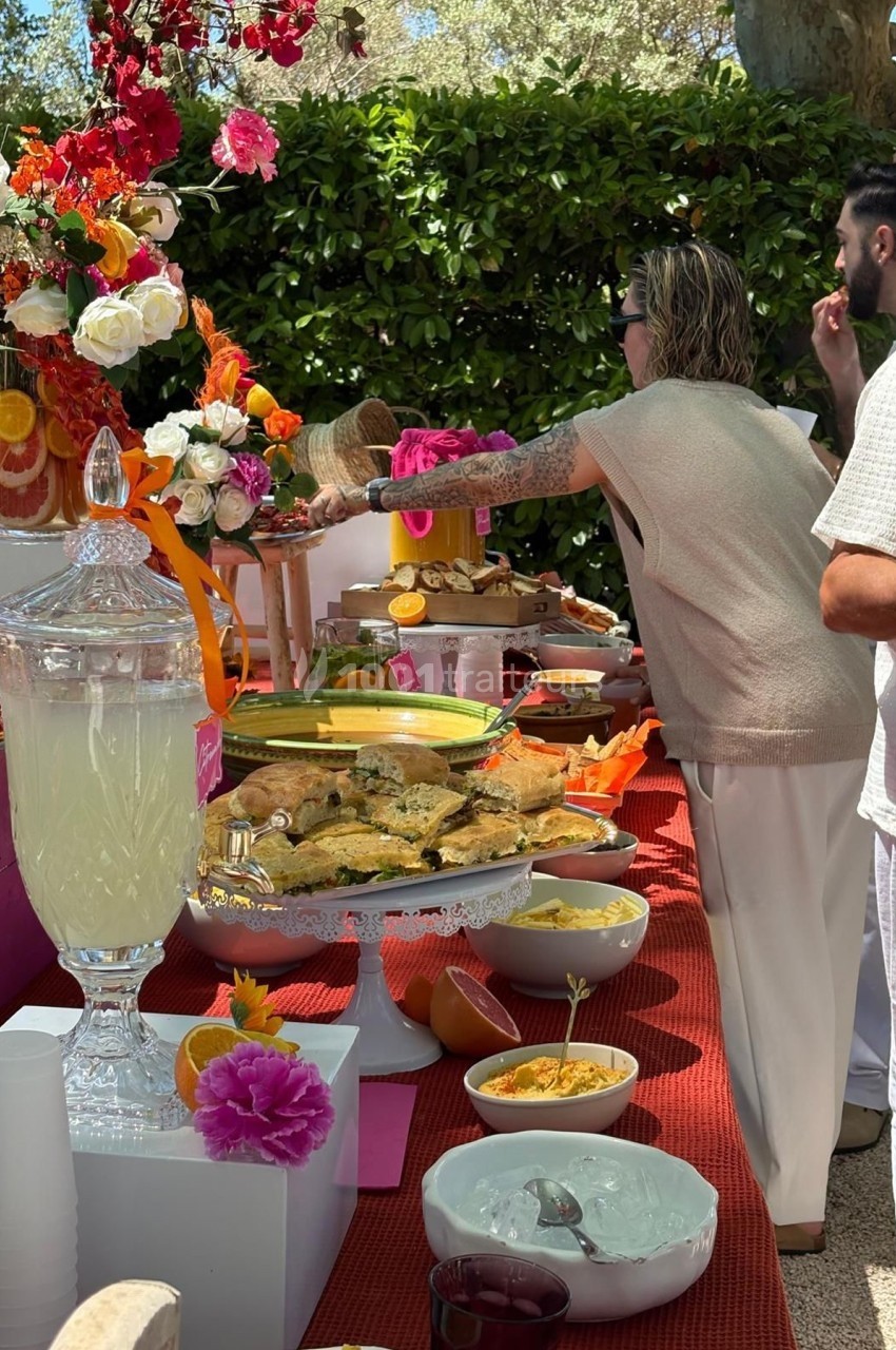 Buffet en plein air avec boissons, plats variés, fleurs colorées et convives se servant.