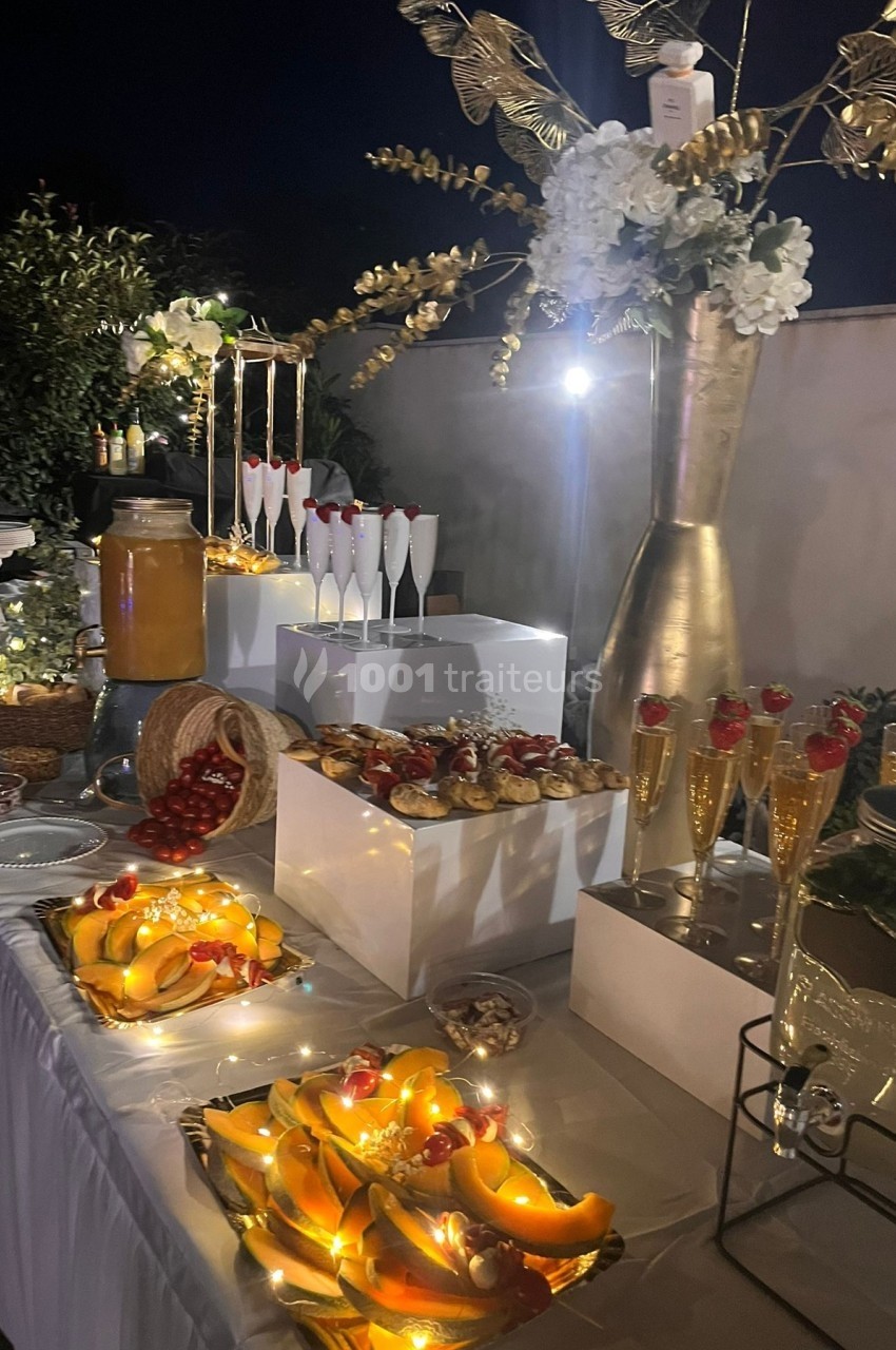 Table de buffet élégamment décorée avec des fruits, pâtisseries, boissons et guirlandes lumineuses, en extérieur nocturne.