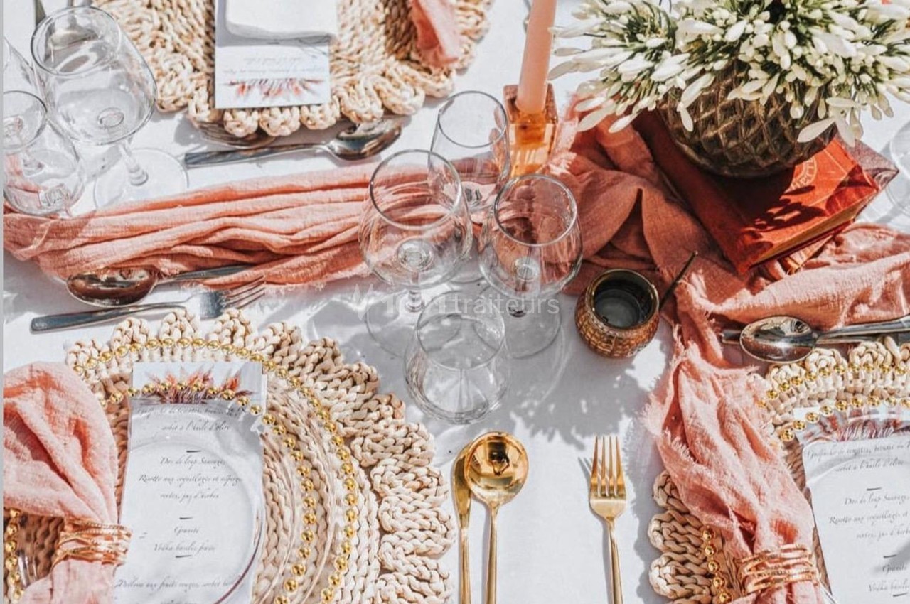 Table dressée avec des assiettes sur des sets en rotin, couverts dorés, serviettes roses et un centre de table floral.