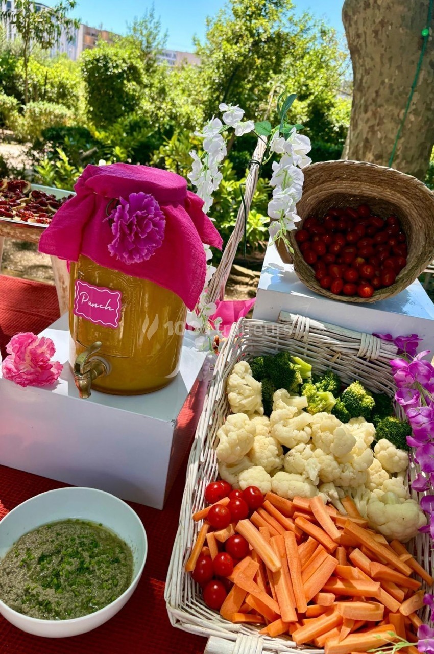 Distributeur de jus décoré, légumes frais en paniers et bol de sauce, disposés sur une table en extérieur.