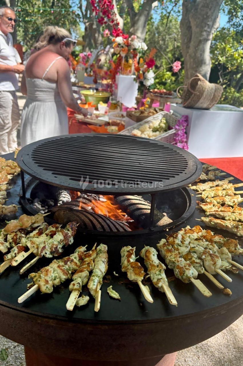 Brochettes de viande grillant sur un barbecue rond lors d'un buffet en plein air, avec des convives en arrière-plan.