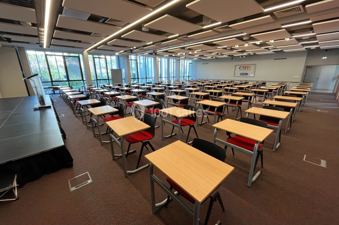Location salle Nice (Alpes-Maritimes) - Campus Sud des Métiers #4 Salle de conférence lumineuse avec des rangées de tables et chaises rouges, équipée d'une scène et de grandes fenêtres.