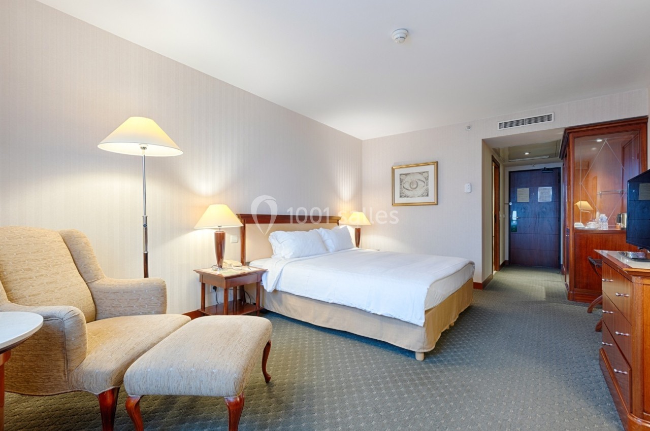 Chambre d'hôtel lumineuse avec lit double, fauteuil, bureau en bois, éclairage doux et porte menant à un couloir.