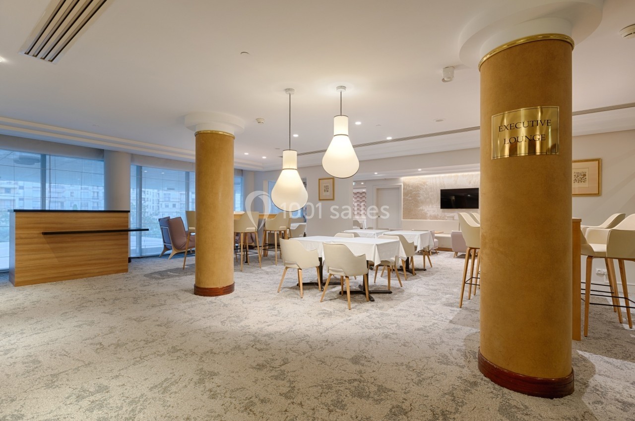 Salle lumineuse avec tables, chaises blanches, colonnes dorées et un comptoir en bois, ambiance moderne et épurée.