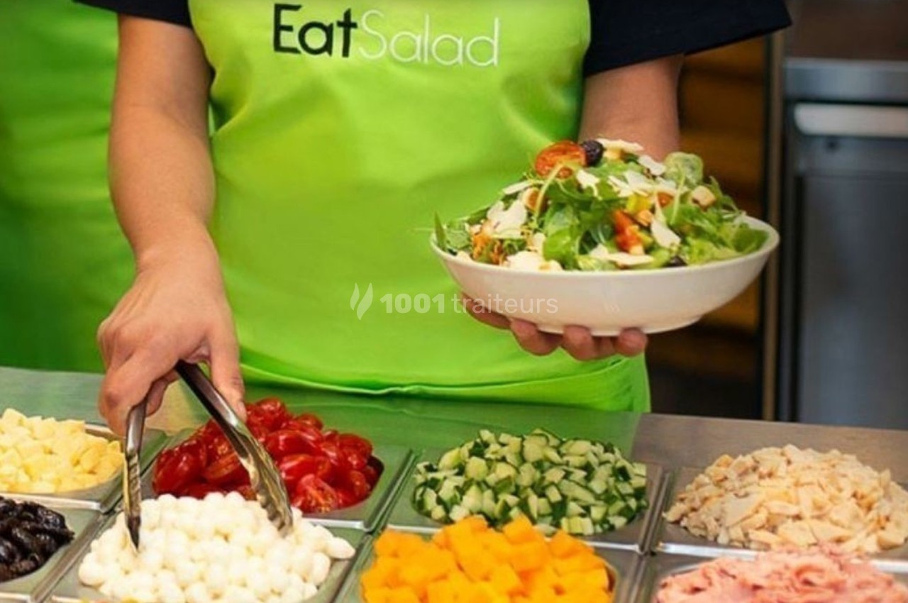 Une personne en tablier vert sert une salade à partir d'un buffet d'ingrédients variés.