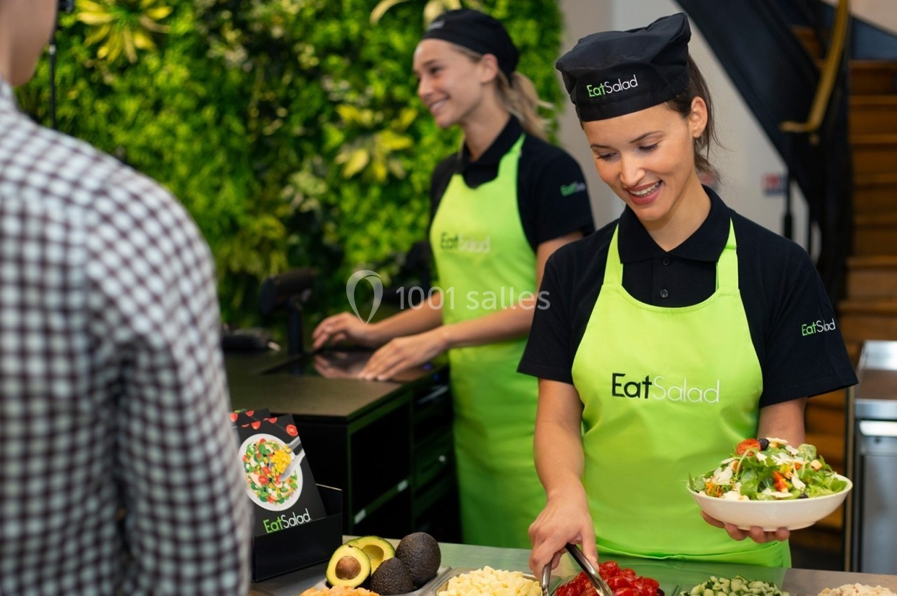 Deux employées en tablier vert préparent des salades fraîches pour un client dans un restaurant lumineux.