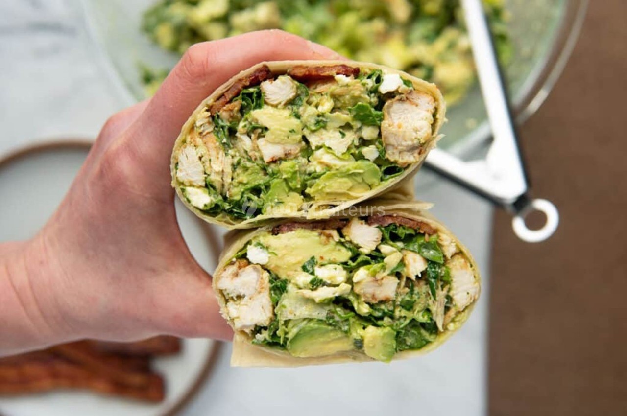 Deux wraps coupés en deux, garnis de poulet, avocat, salade et autres ingrédients frais, tenus dans une main.