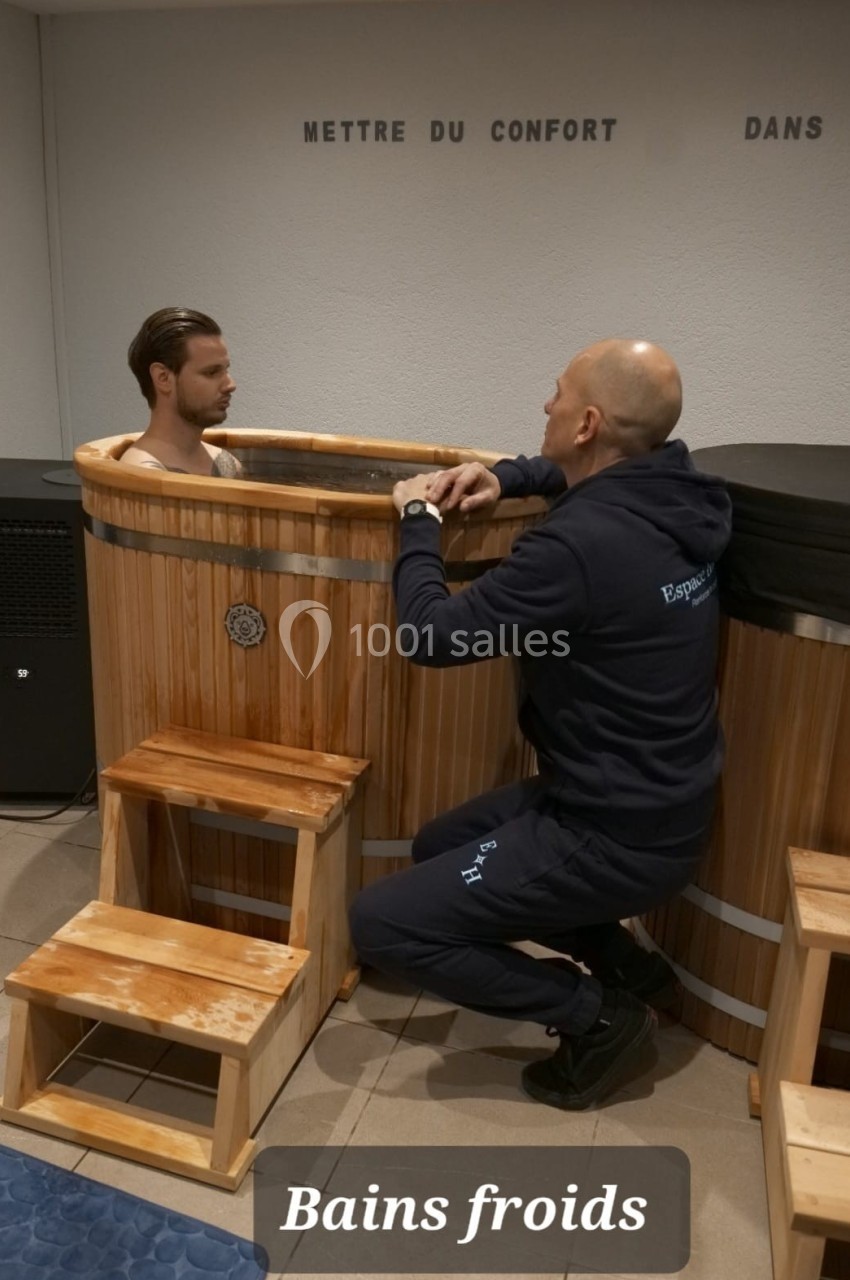 Un homme immergé dans un bain froid en bois, accompagné d'un autre homme qui l'encadre.