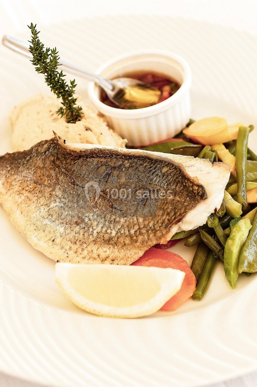 Filet de poisson grillé servi avec légumes, purée, citron et une sauce dans un ramequin blanc.