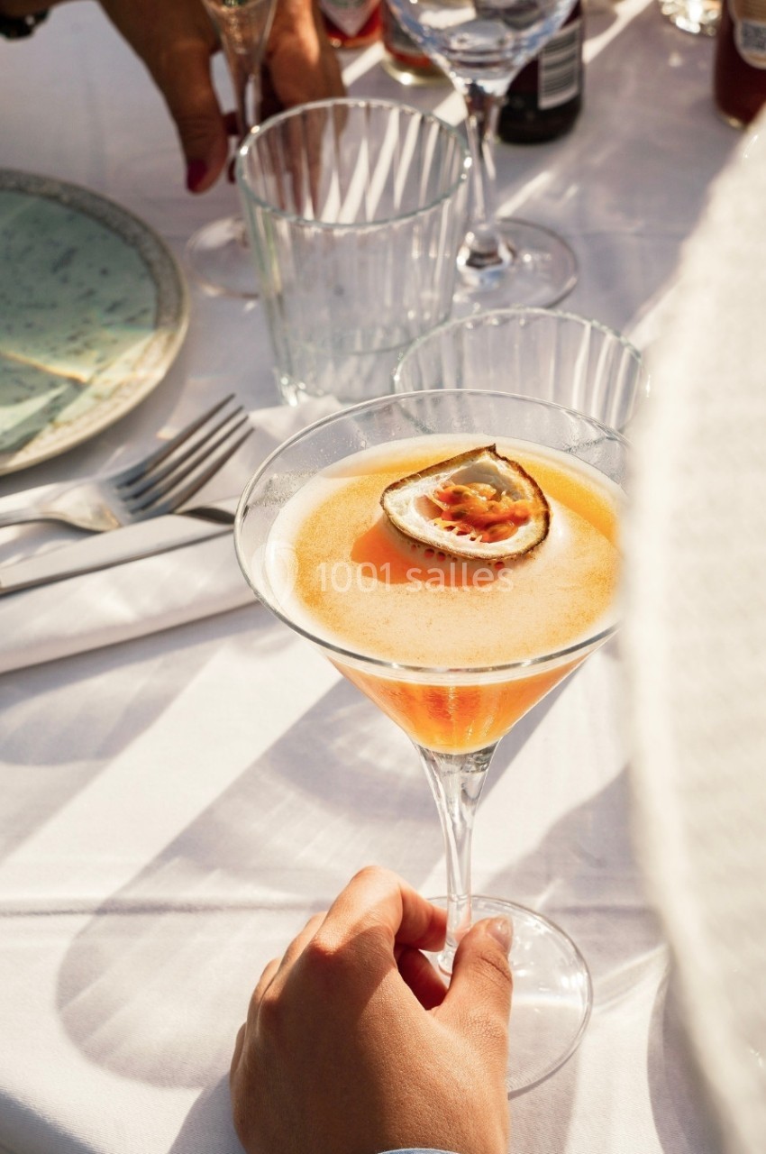 Un verre à cocktail garni d'un demi-fruit de la passion, posé sur une table dressée en extérieur.