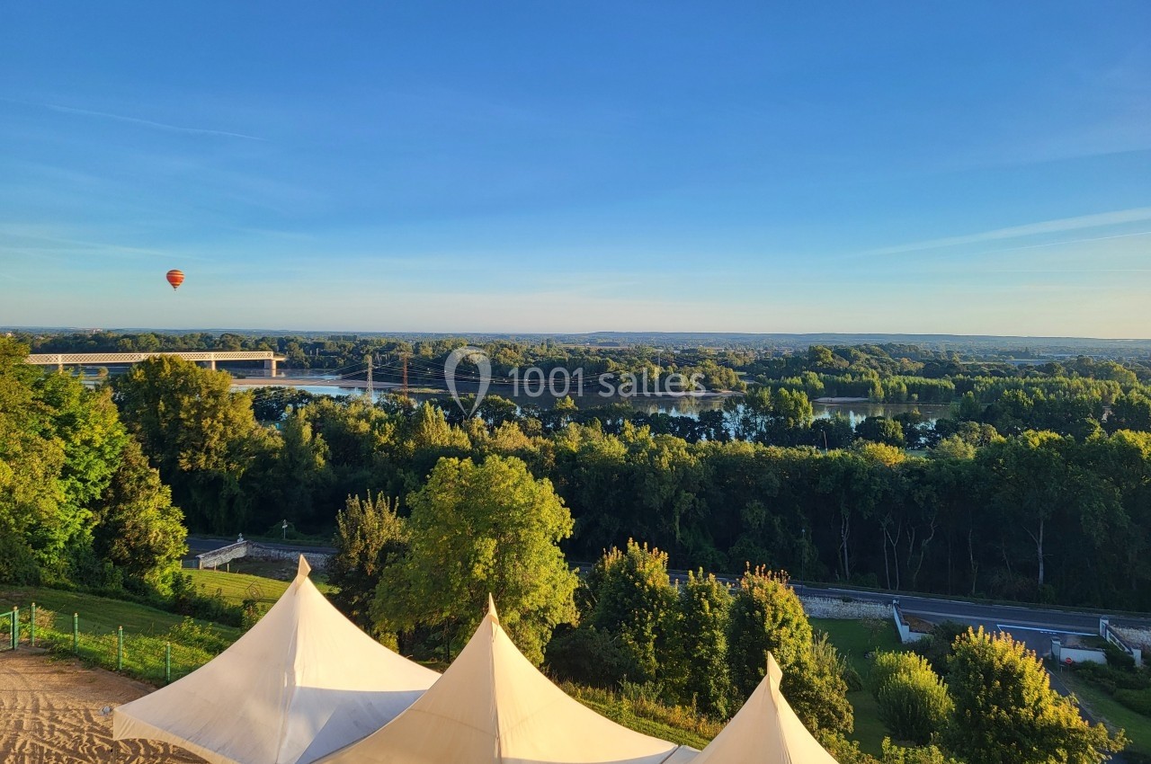 Paysage avec des tentes blanches au premier plan, une rivière, un pont et une montgolfière sous un ciel dégagé.