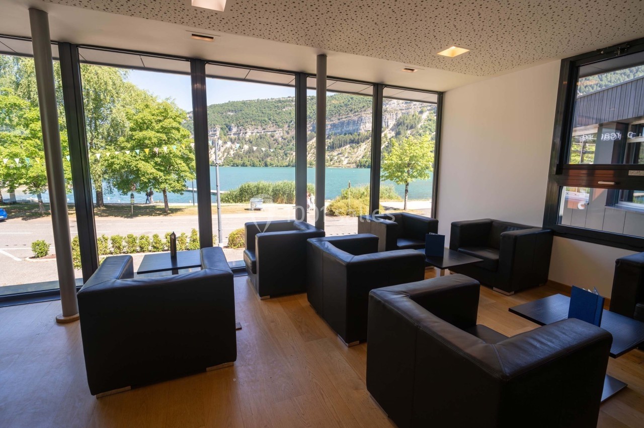 Salon avec fauteuils noirs, grandes baies vitrées offrant une vue sur un lac, des arbres et des collines verdoyantes.