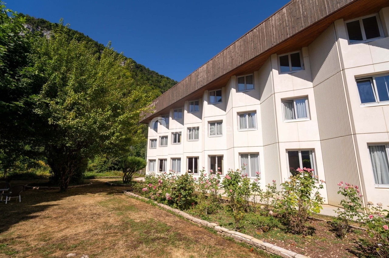 Façade d'un bâtiment moderne entouré de rosiers et d'arbres, avec un ciel dégagé en arrière-plan.