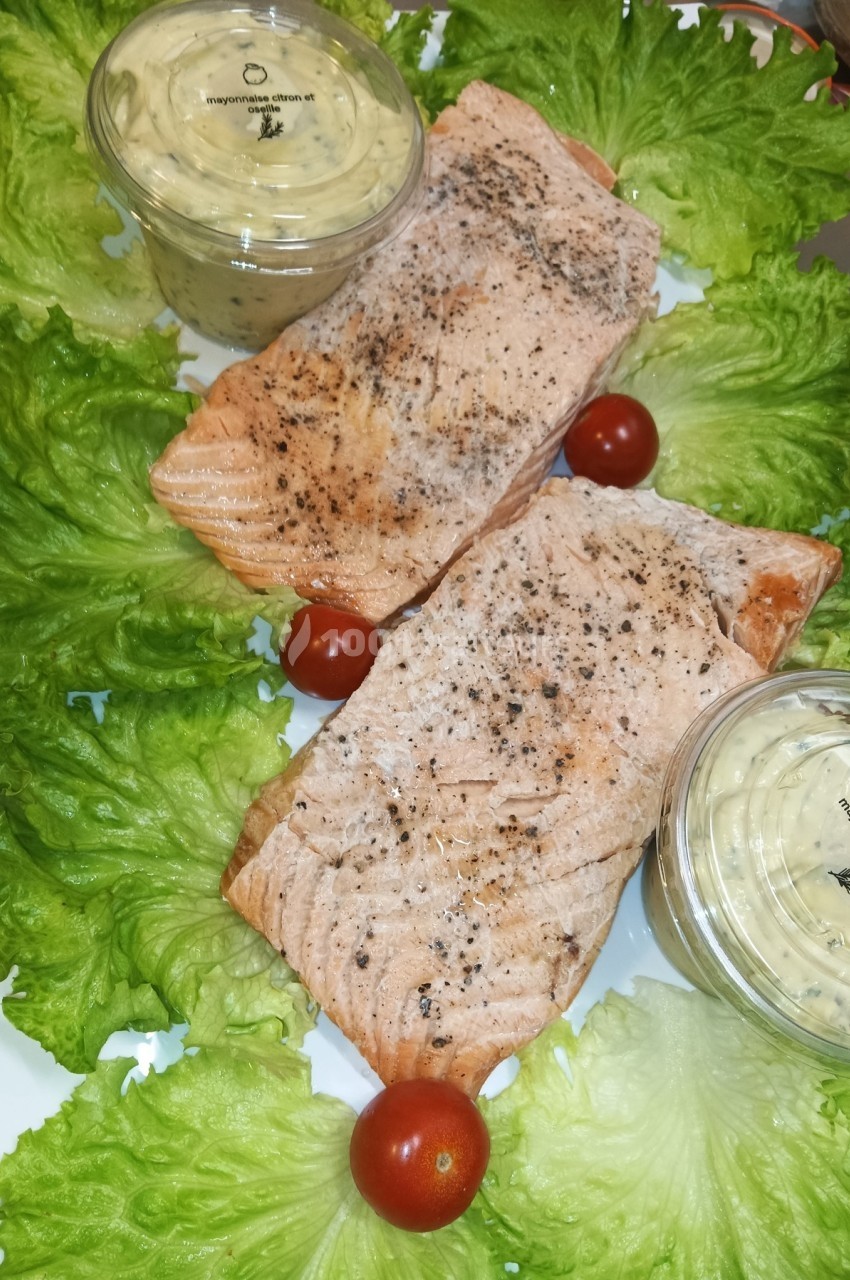 Deux filets de saumon cuits, assaisonnés, disposés sur un lit de laitue avec des tomates cerises et des sauces en pot.