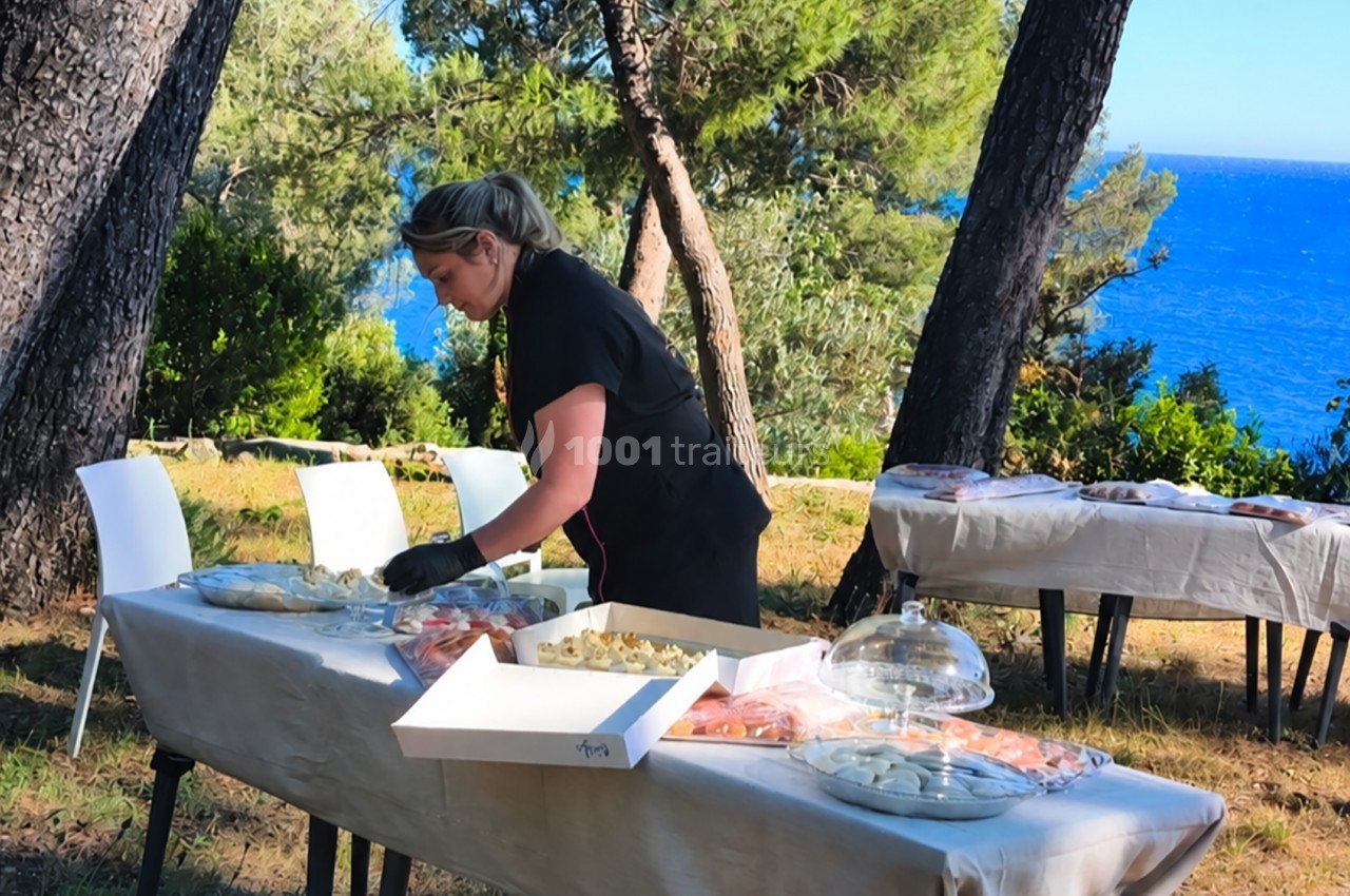 Une femme prépare un buffet en plein air près de la mer, entourée d'arbres et de tables dressées.