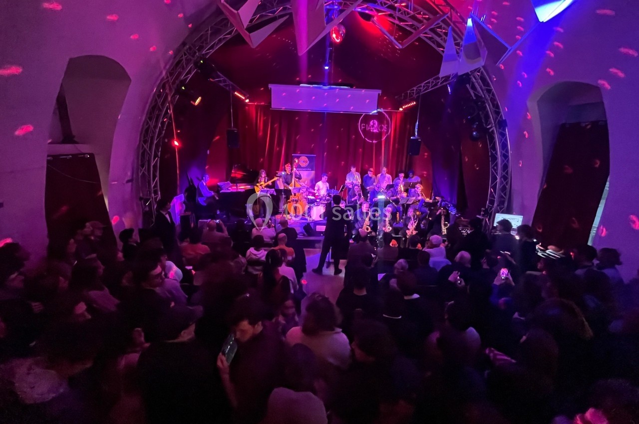 Concert en intérieur avec un groupe sur scène, éclairages colorés et public nombreux dans une ambiance festive.