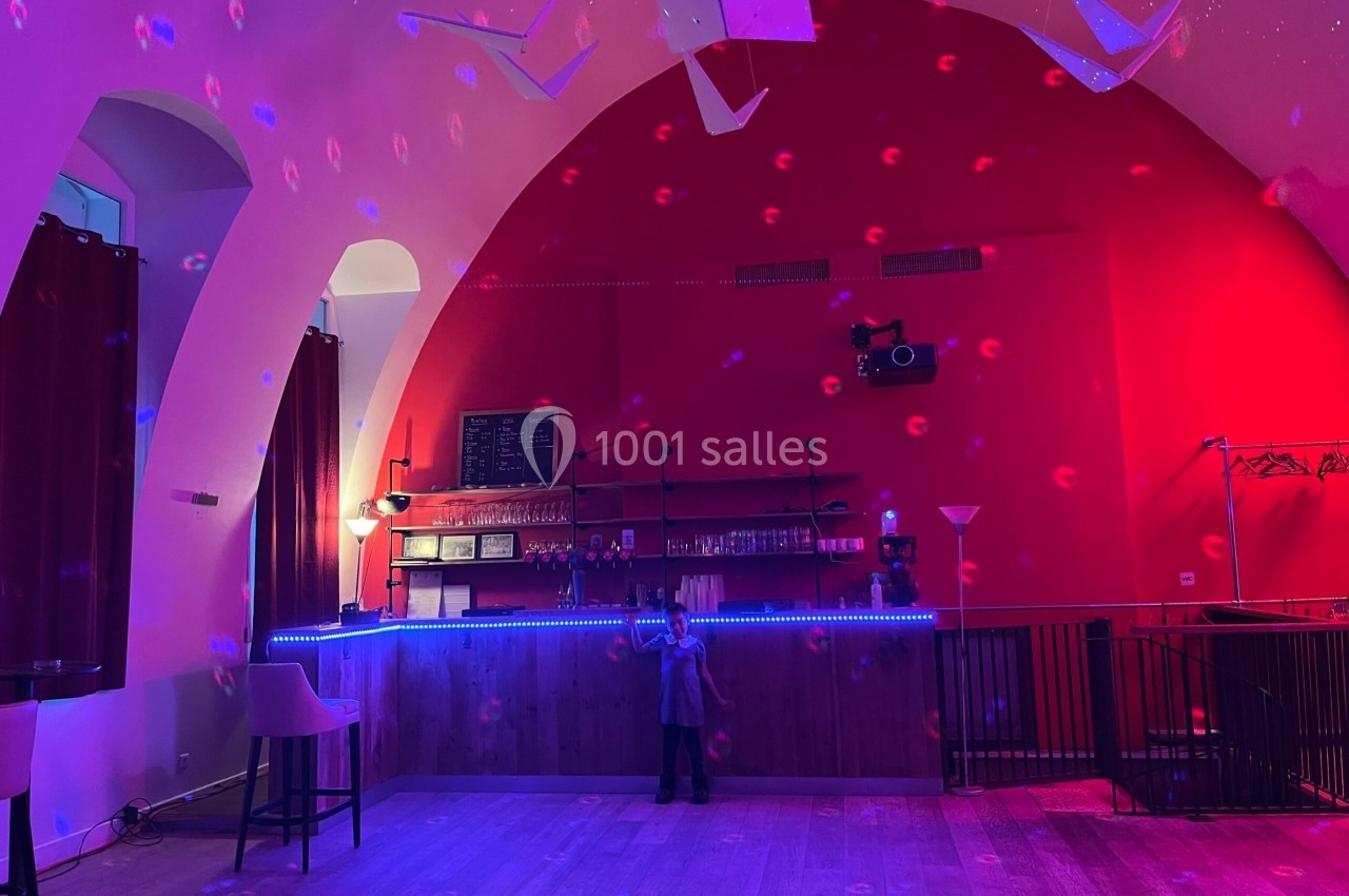 Salle avec un bar éclairé par des lumières colorées, un enfant debout au centre sous un plafond voûté rouge.